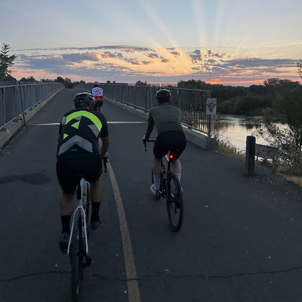 Two cyclists ride on a bridge during sunset, with a river and trees visible in the background. The cyclist in the foreground wears a black and neon yellow-green jersey, and the scene is lit by warm sunset light with dramatic sun rays.
